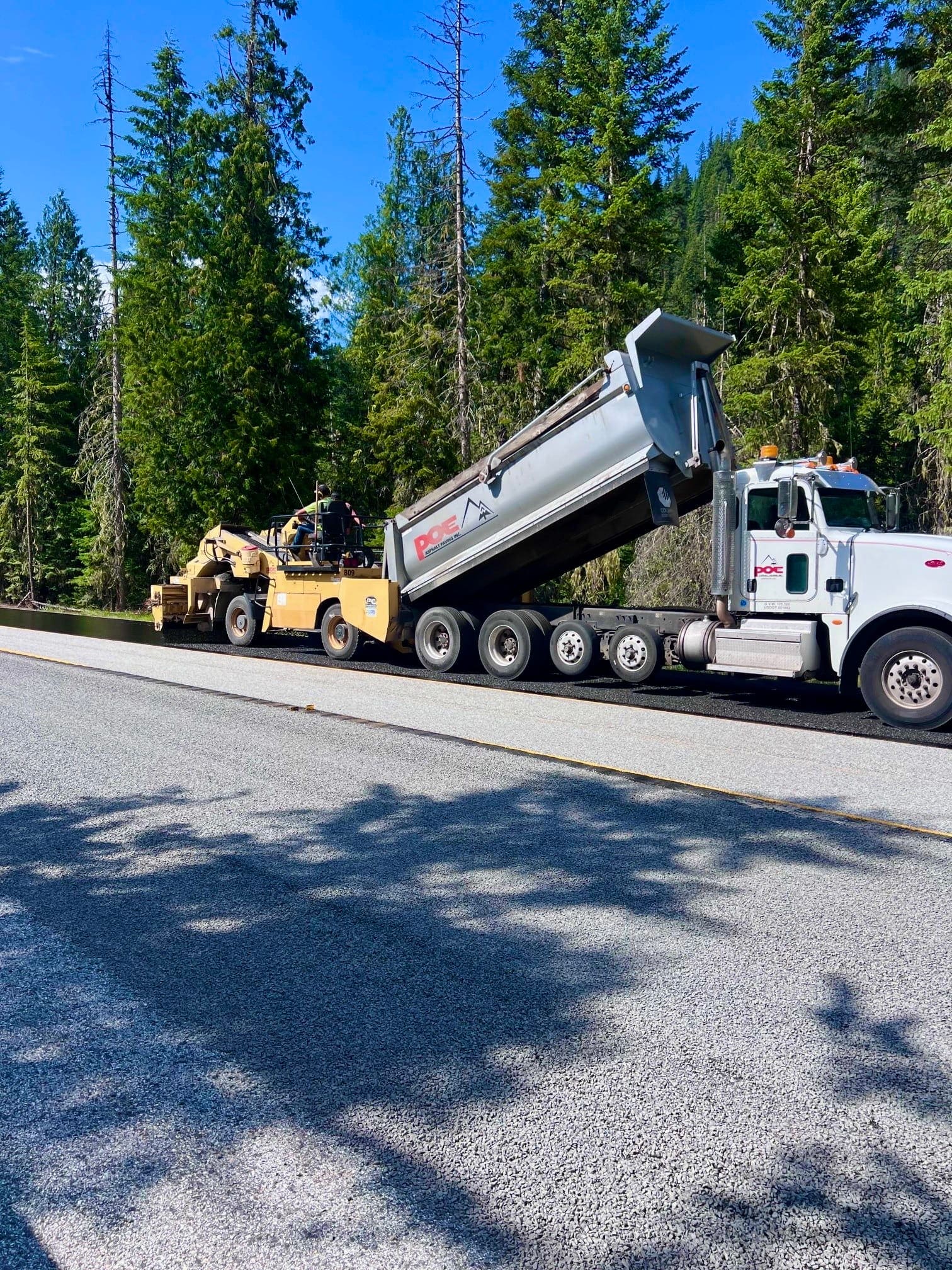 Truck unloading asphalt into road paving machine on forest-lined highway under clear blue sky.