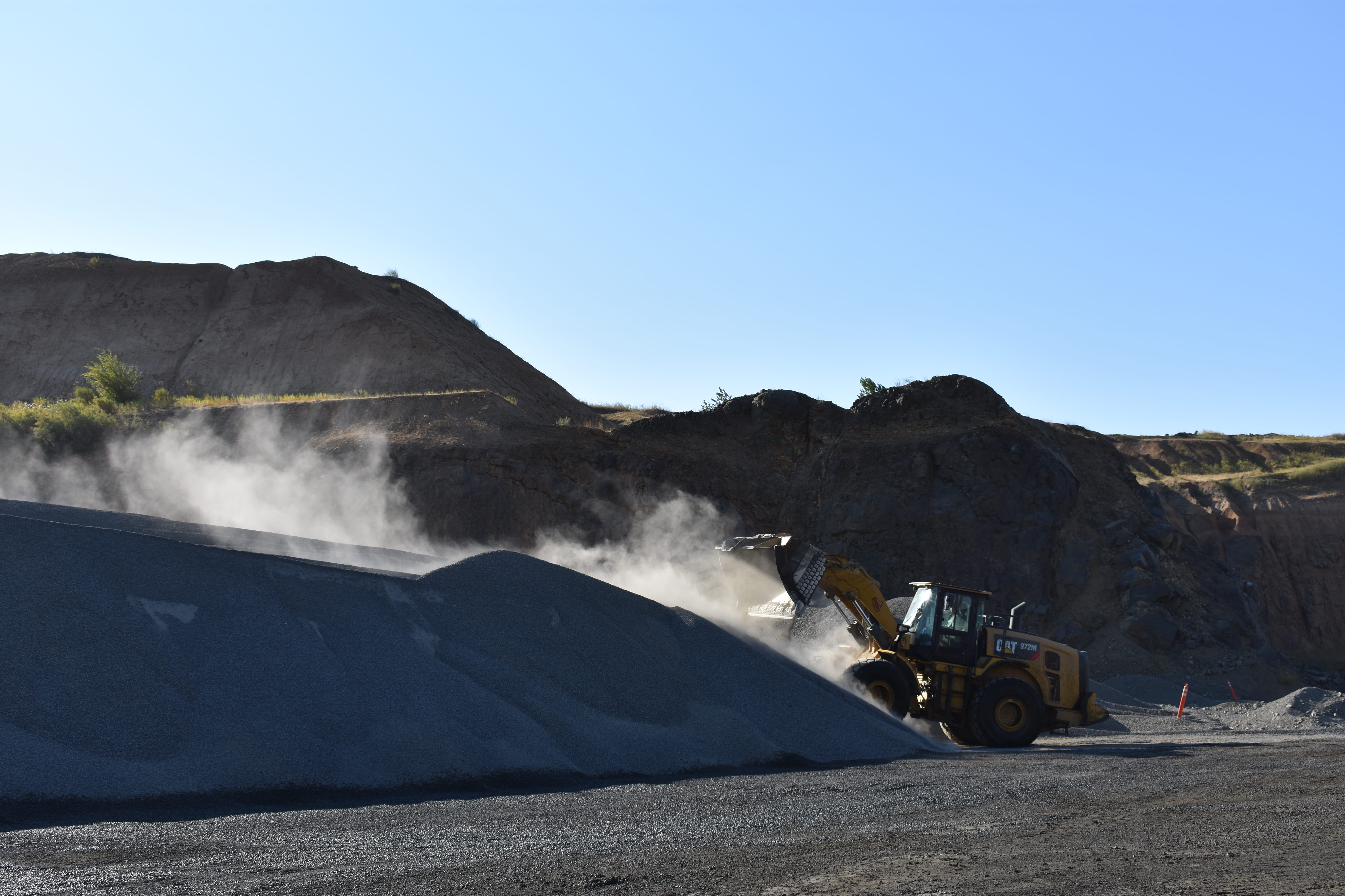 Yellow loader dumping gravel onto a large pile at a quarry with dust cloud and rocky hills in background.x