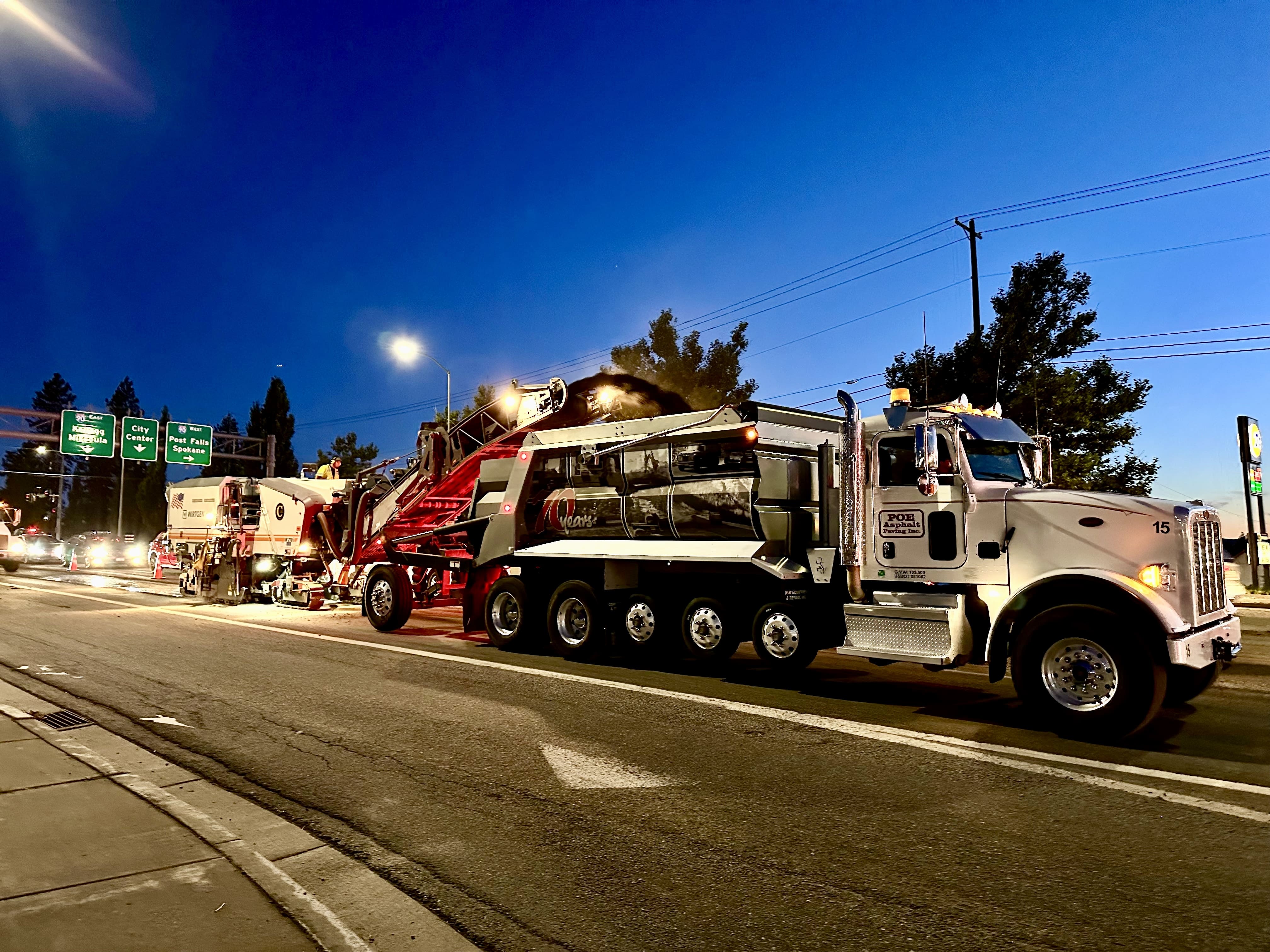Asphalt paving truck and machinery working on a city street at dusk under streetlights.