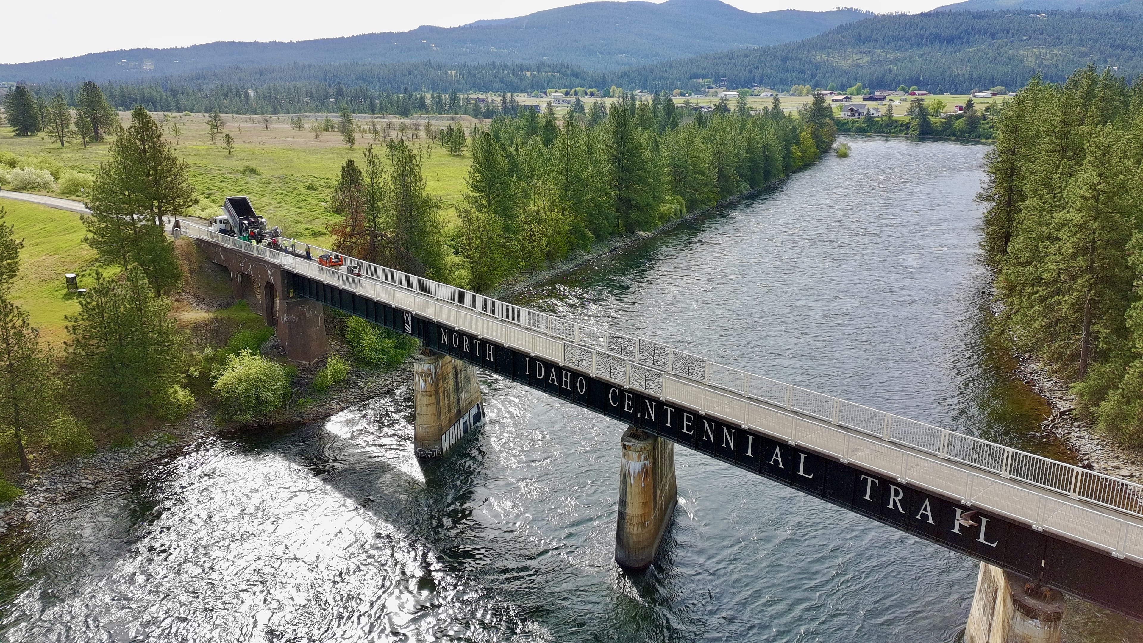 Bridge over river labeled North Idaho Centennial Trail with green trees and hills in the background.