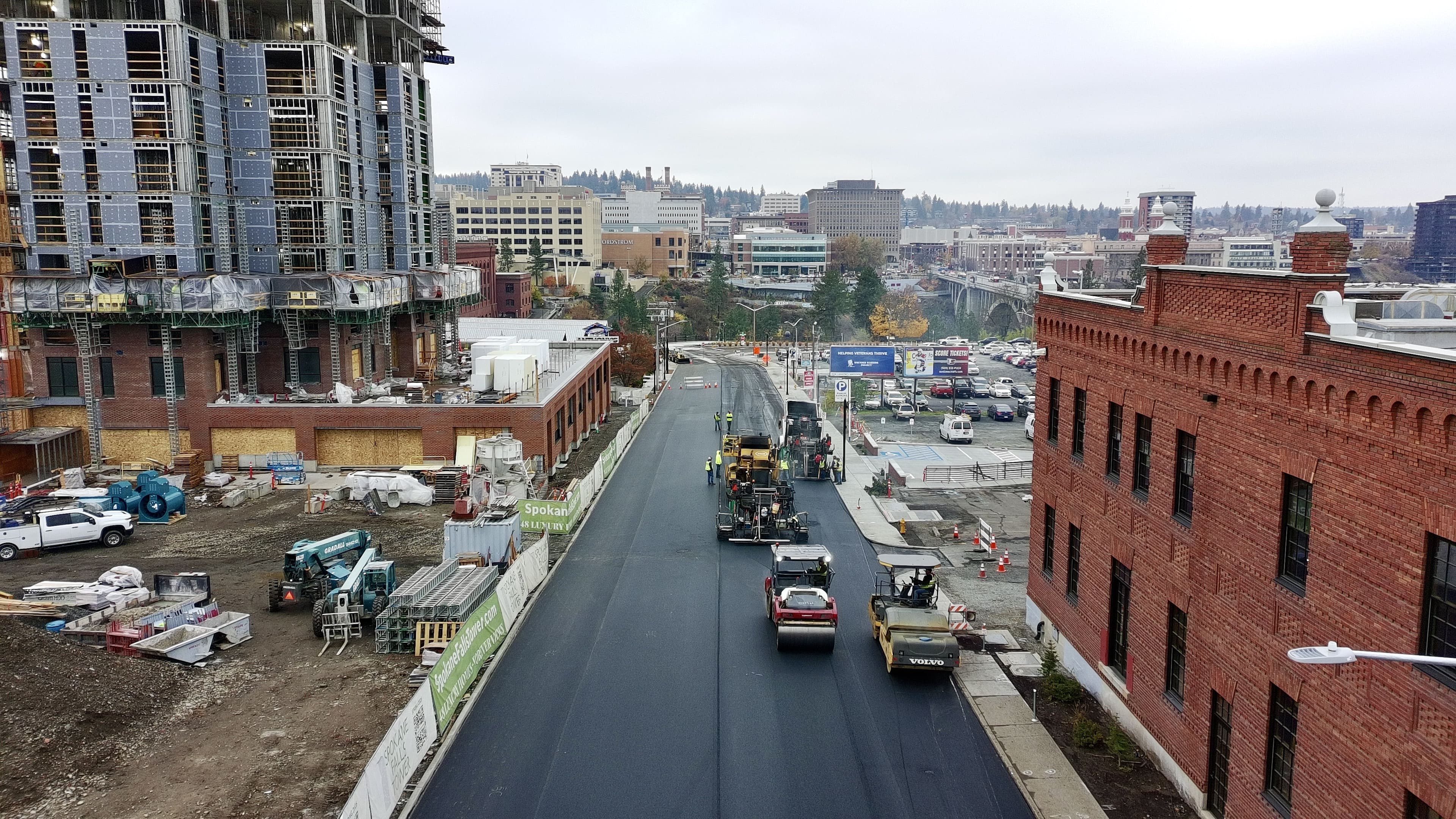 Construction workers paving a new asphalt road between buildings in an urban area under overcast sky.