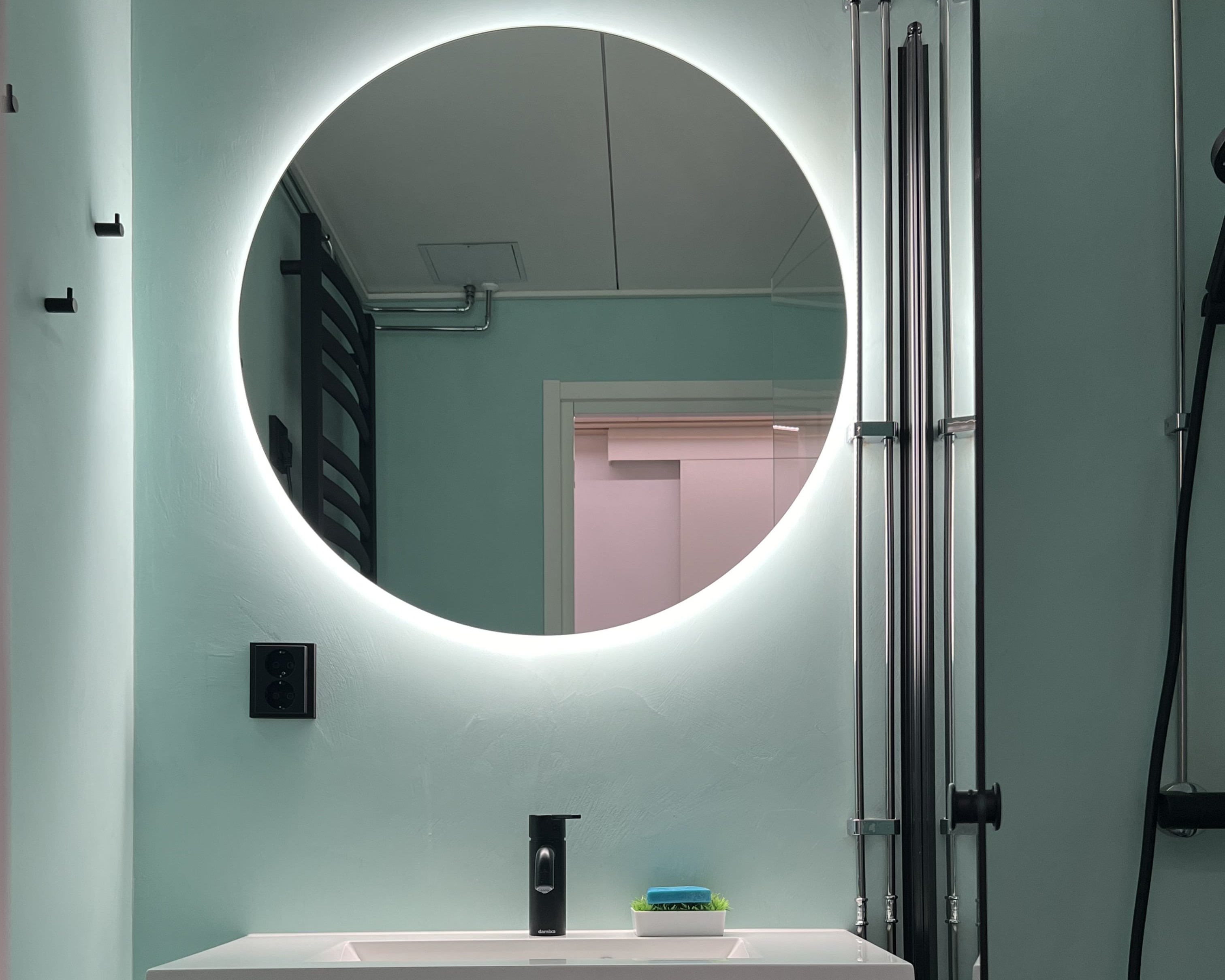 Pyöreä, taustavalaistu peili vaaleansinisessä kylpyhuoneessa, jossa on musta hana ja saippua-alusta.