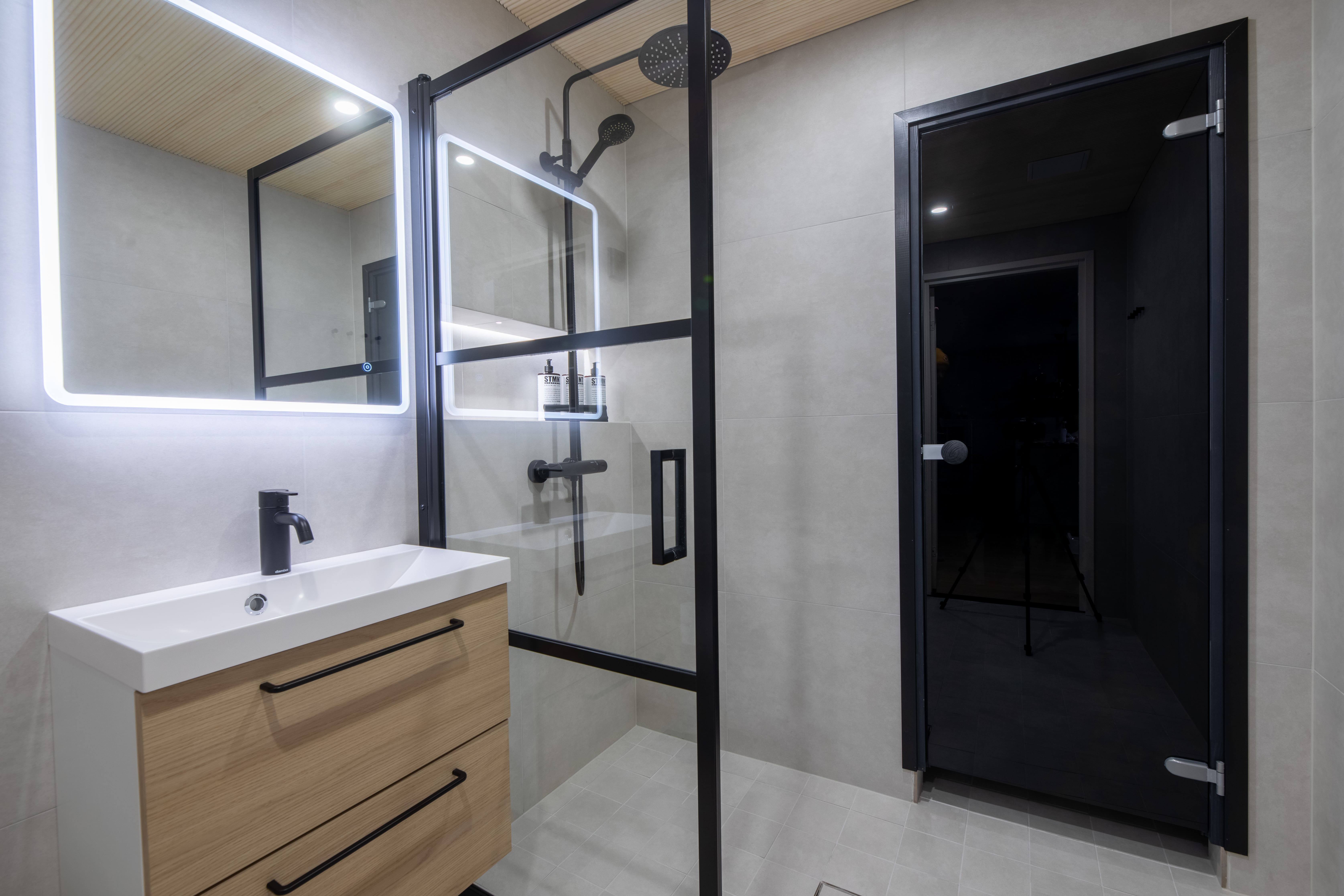 Modern bathroom with a wooden vanity, LED-lit mirror, glass shower enclosure with black fixtures, and a dark sauna door.