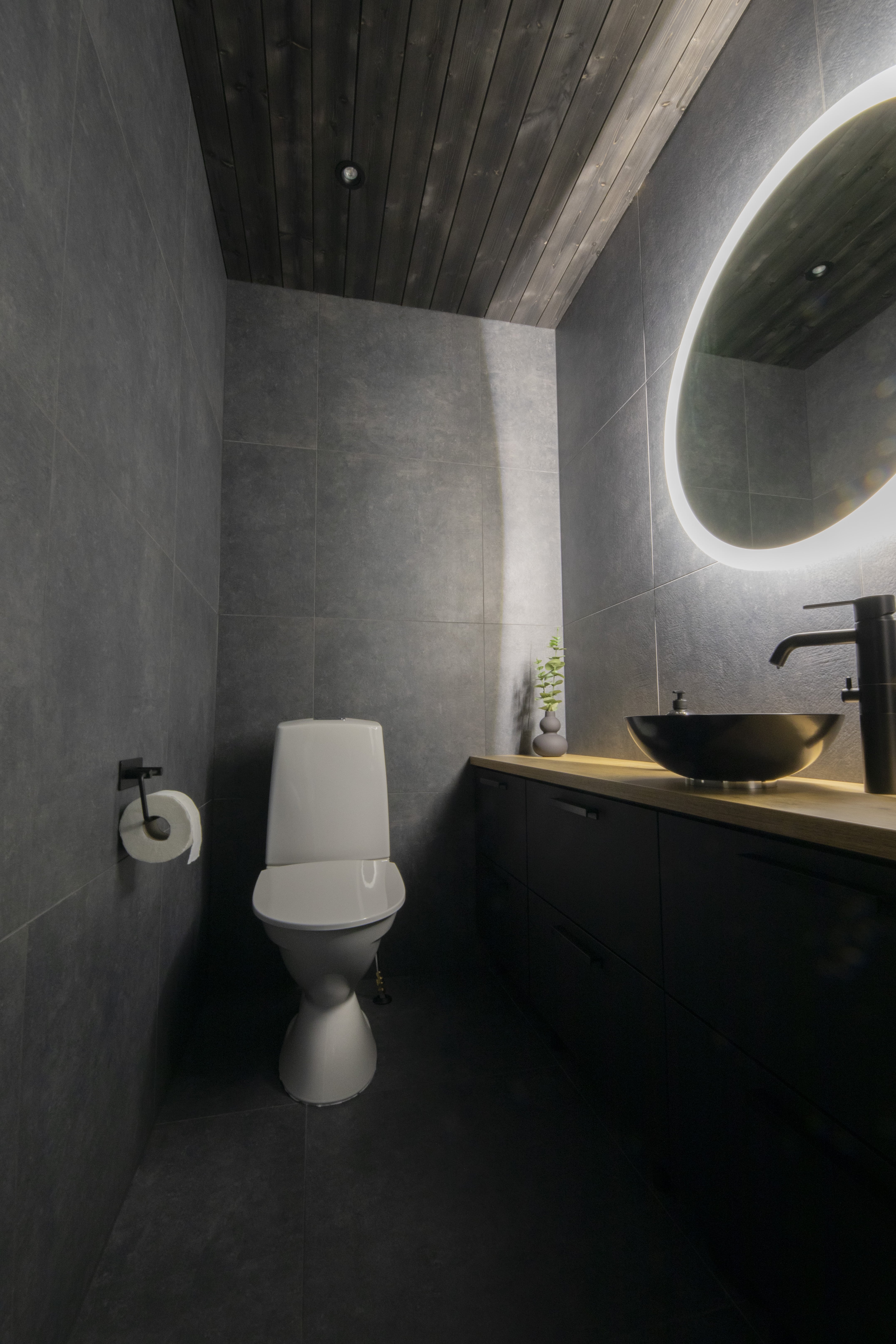 Tyylikäs moderni kylpyhuone, jossa valkoinen wc-istuin, musta pesuallas, pyöreä valaistu peili ja puinen katto.