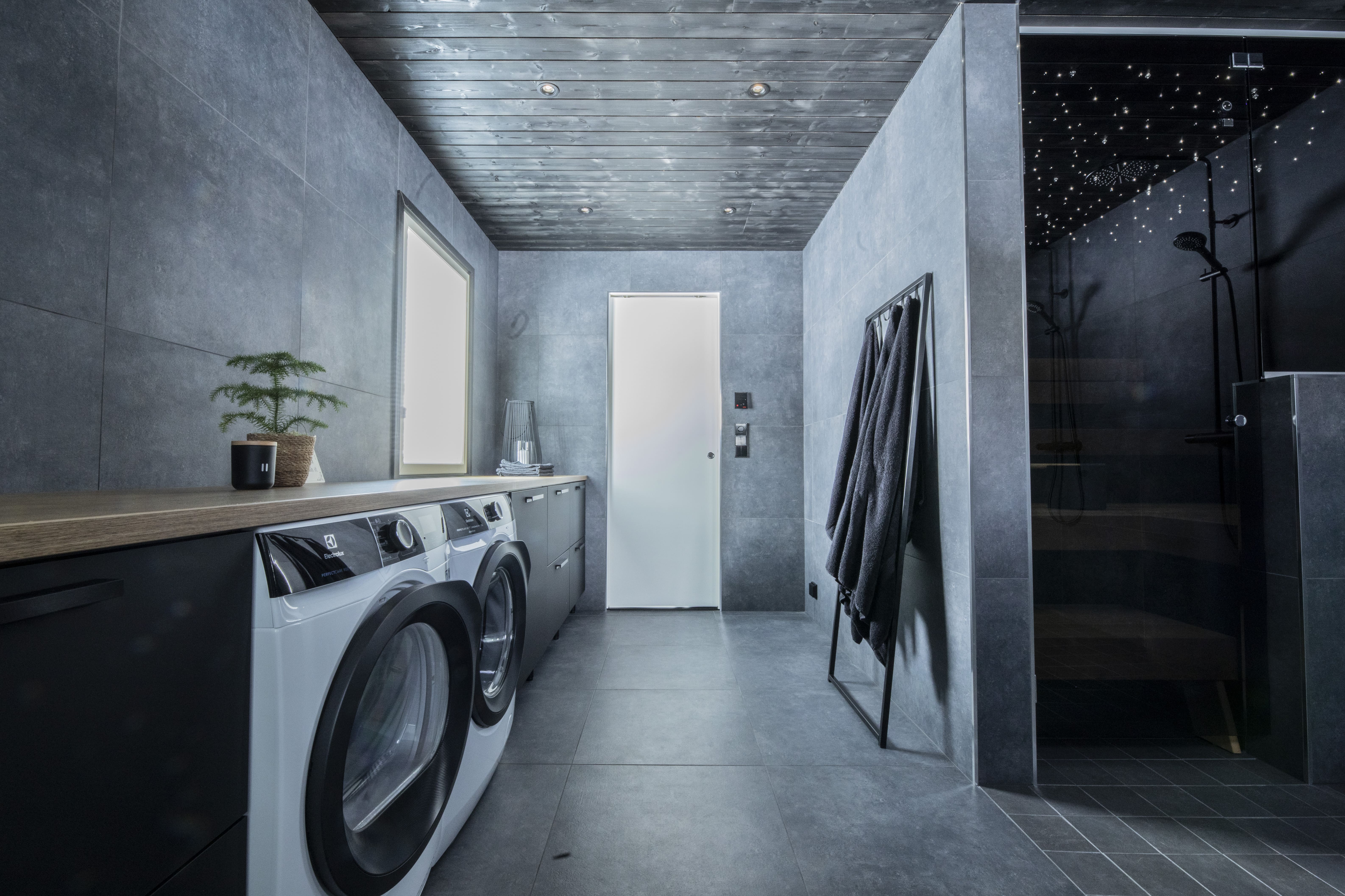 Moderni pesuhuone, jossa on pyykinpesukone ja kuivausrumpu, puiset kaapit, harmaat laatat ja musta suihkutila tähtivaloilla.