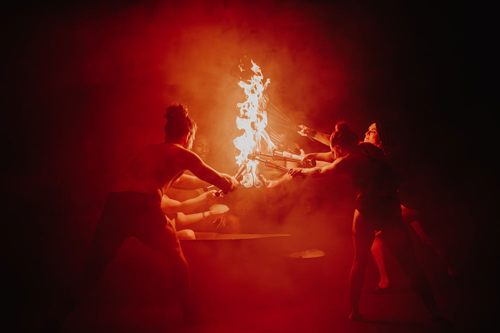 Group of performers engaging in fire dancing with torches and flames, surrounded by smoke and red lighting.