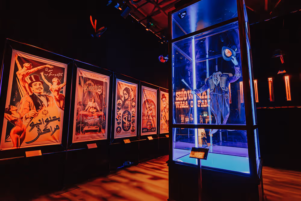 Dimly lit museum exhibit with vintage circus posters on the wall and a suspended costume displayed in a glass case illuminated with blue light.