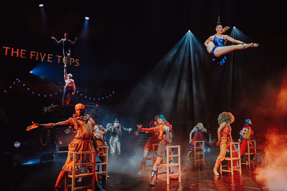 Performers in colorful costumes on stage with chairs, including two acrobats balancing vertically and a woman suspended mid-air in a blue outfit under spotlights.