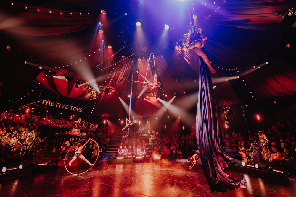Circus performers executing aerial silks and acrobatics on a stage with a seated audience under dramatic red lighting.