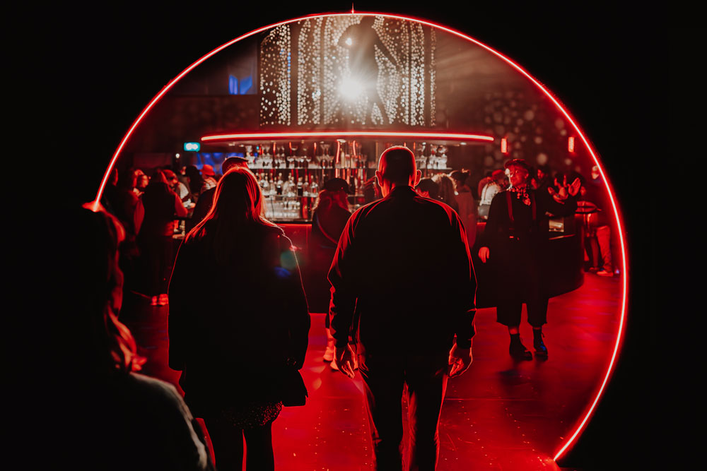 People standing and walking inside a bar area illuminated by a red neon circular light frame.