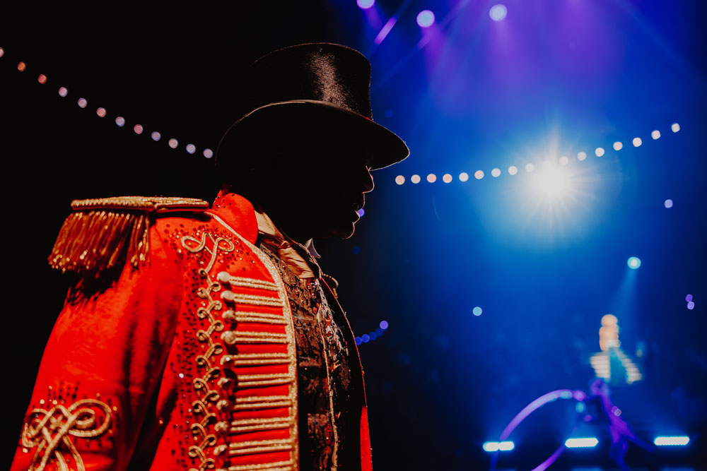 Silhouette of ringmaster wearing a red, ornate ringmaster costume and black top hat under stage lights with a blurred audience background.