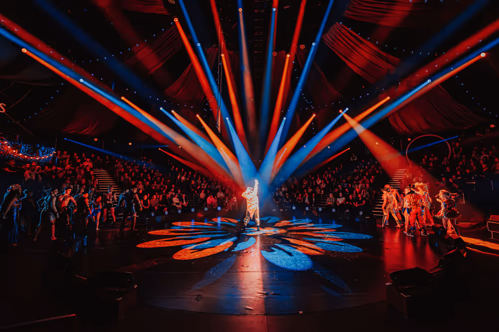 Performers standing on a stage illuminated by dramatic red and blue spotlights with a large audience seated in the background.
