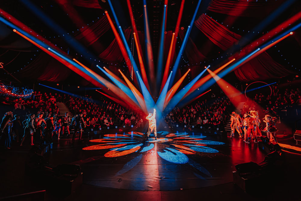 Performers standing on a stage illuminated by dramatic red and blue spotlights with a large audience seated in the background.