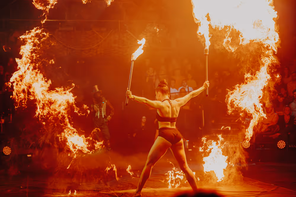 Performer in black outfit holding flaming torches with large fire arcs around her on stage in front of an audience.