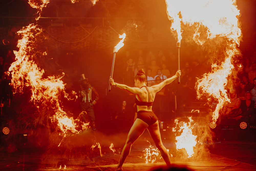 Performer in black outfit holding flaming torches with large fire arcs around her on stage in front of an audience.