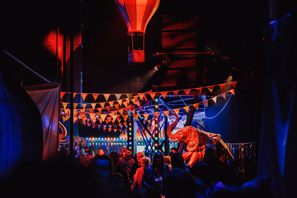 Crowd walking under red and white pennant flags with a red and white hot air balloon and an illuminated elephant figure in a dark, festive indoor space.