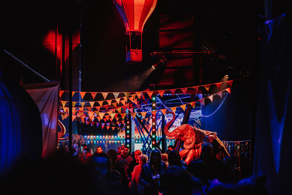 Crowd walking under red and white pennant flags with a red and white hot air balloon and an illuminated elephant figure in a dark, festive indoor space.