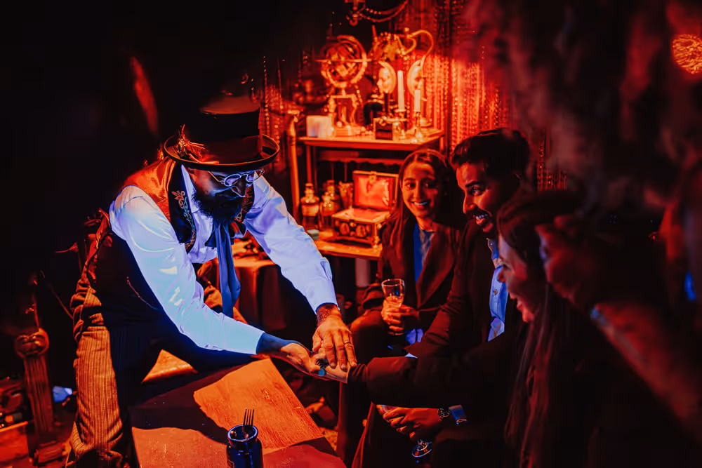 Magician in vintage costume performing a close-up card trick for a group of smiling people in a dimly lit room with warm red lighting.