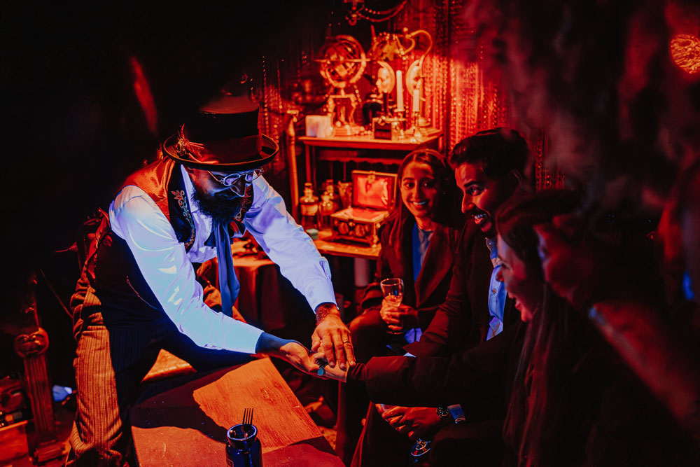 Magician in vintage costume performing a close-up card trick for a group of smiling people in a dimly lit room with warm red lighting.