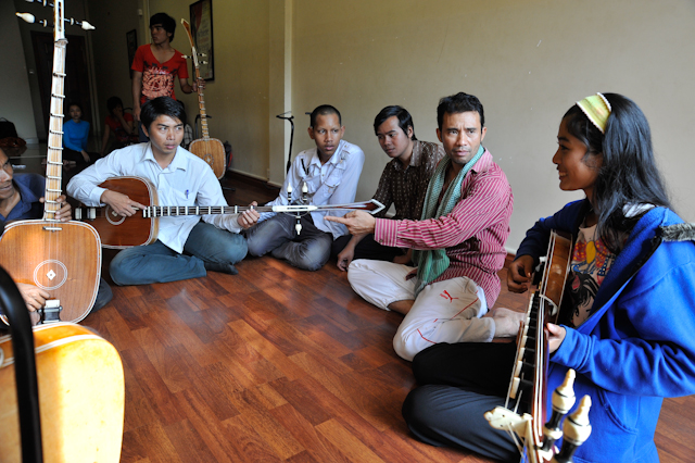 Group of six people sitting on a wooden floor inside a room, some holding guitars and musical instruments.