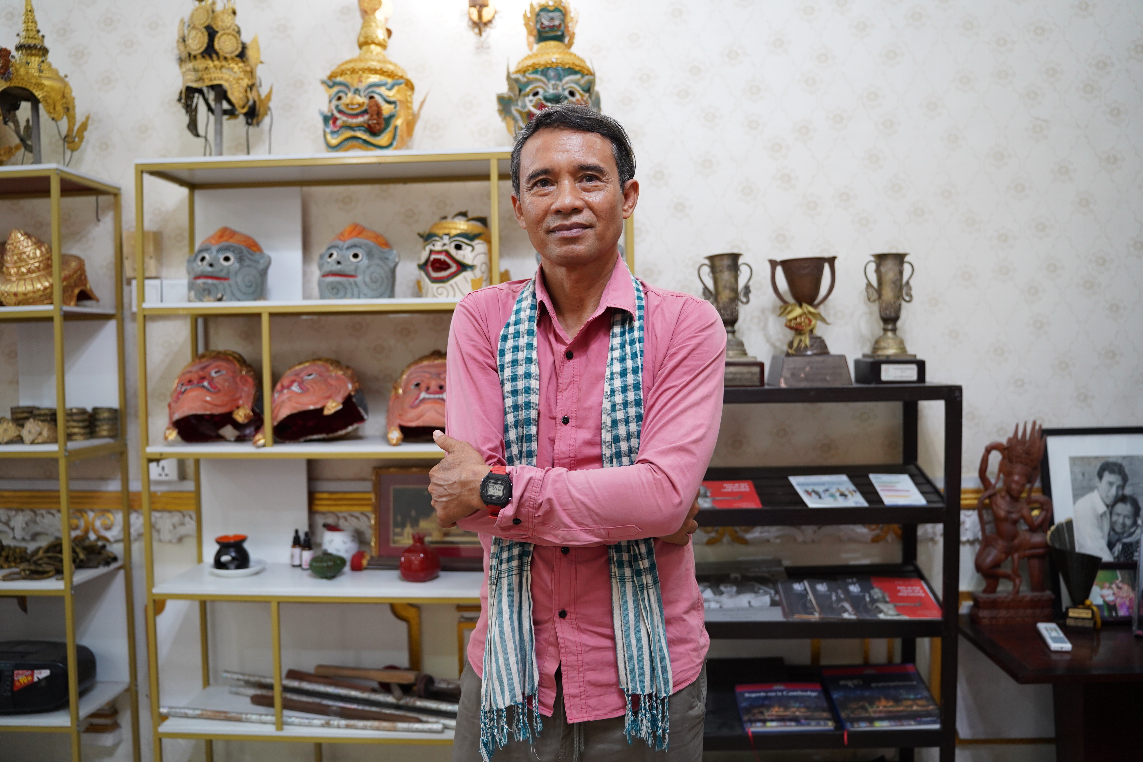 Man in a pink shirt and checkered scarf standing with arms crossed in a room filled with traditional masks, trophies, and framed photos on shelves.