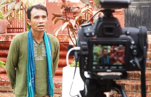 Man in green shirt and blue scarf speaking in front of a camera outdoors with red brick stairs and plants in the background.