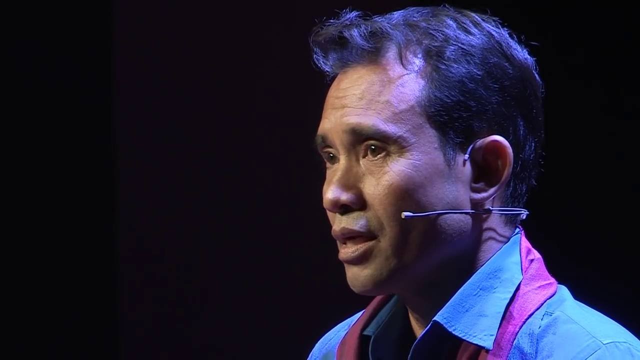 Man wearing glasses and a colorful checkered shirt with a pink scarf speaking at a podium with a microphone and water glass.