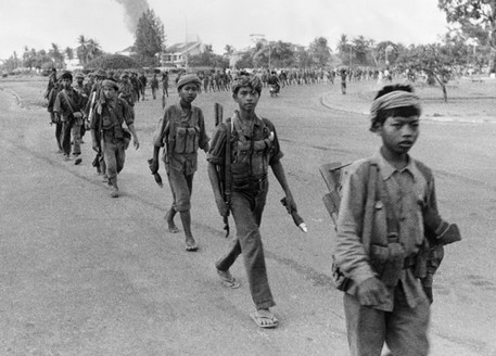 Black and white photo of a line of young soldiers walking barefoot on a curved road, carrying rifles and wearing military gear.
