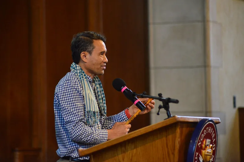 Man in checkered shirt and scarf speaking at a wooden podium with microphone indoors.