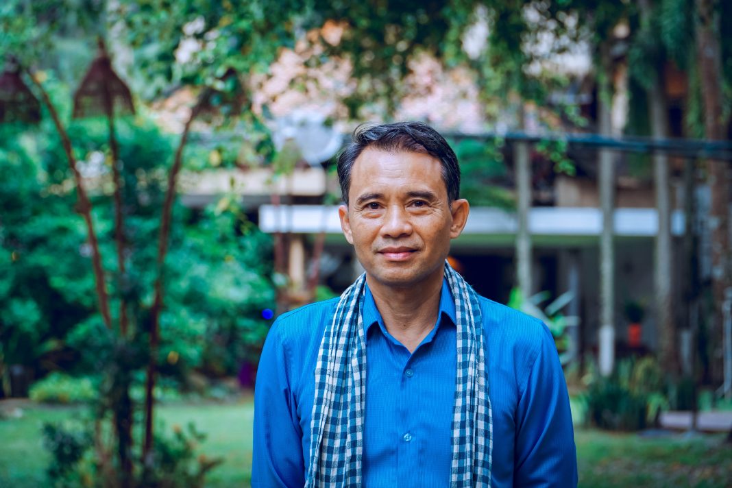 Man in blue shirt with checkered scarf standing outdoors with green trees in the background.