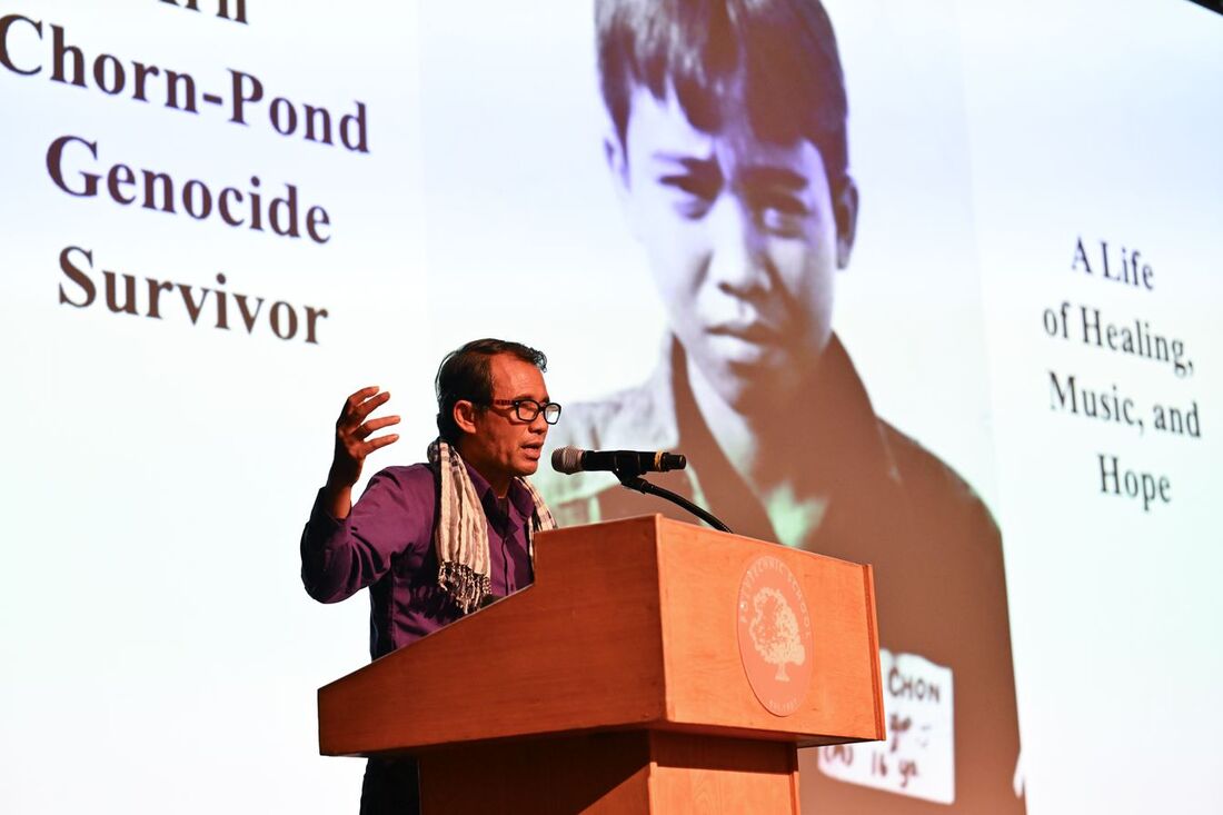 Speaker addressing an audience at a podium with a large projection behind him displaying text about a Chorn-Pond genocide survivor and a black-and-white portrait.