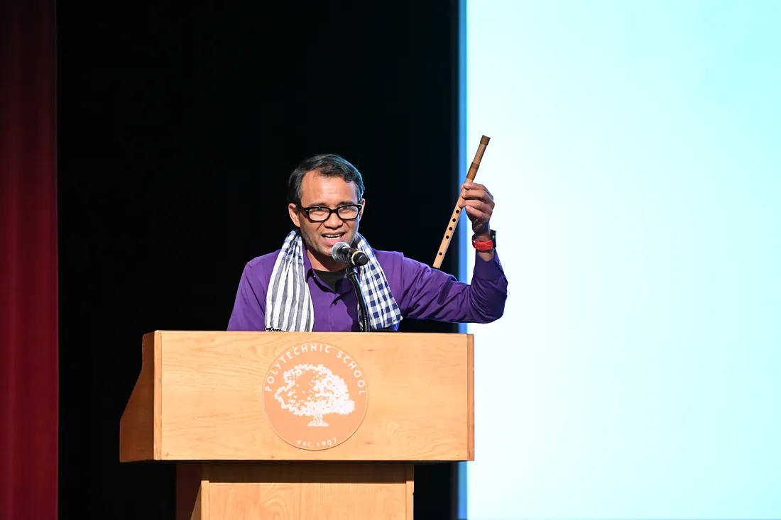 Man wearing glasses and a purple shirt speaks at a wooden podium with a microphone while holding a flute.