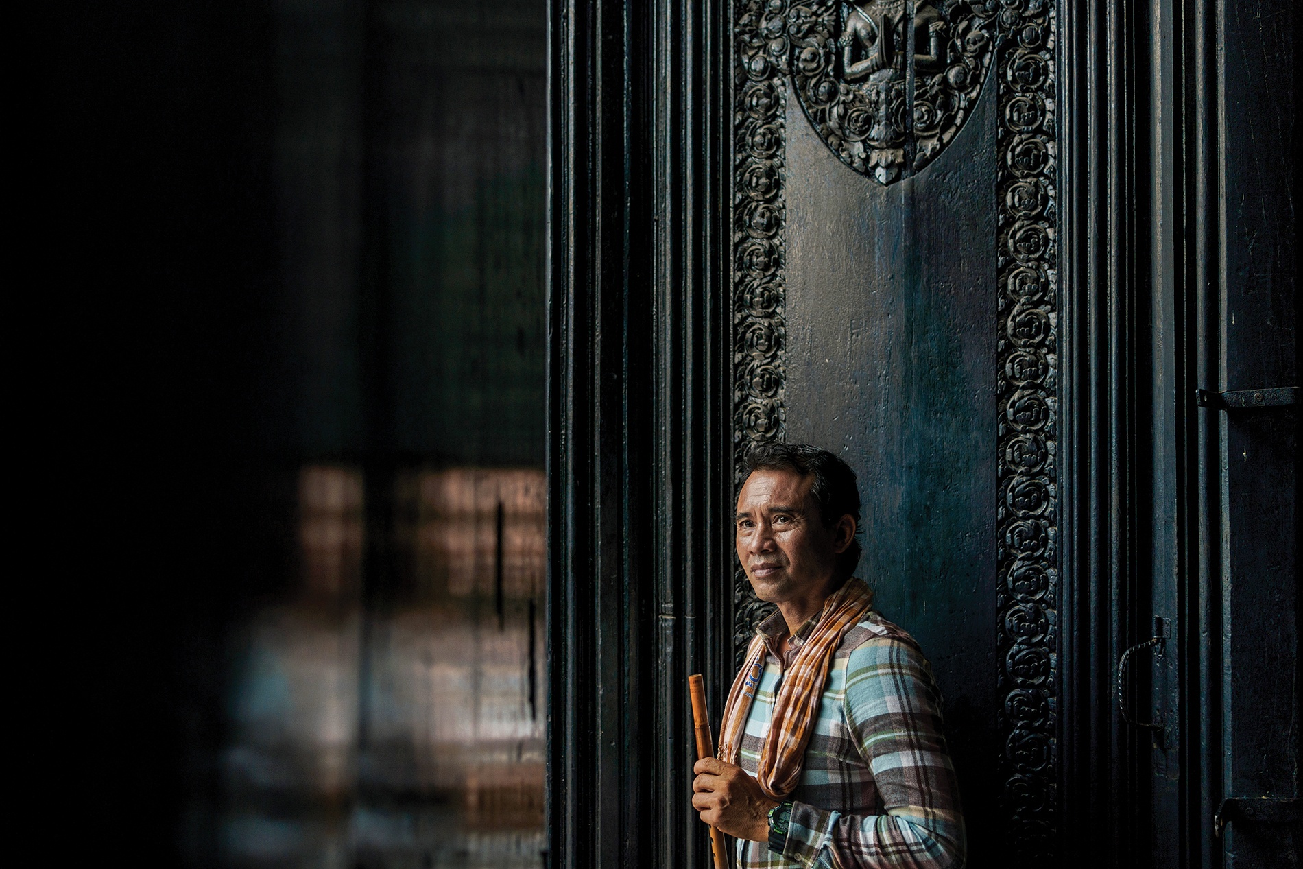 Man in plaid shirt and orange scarf holding a wooden flute, standing by a dark ornate door, gazing thoughtfully to the side.