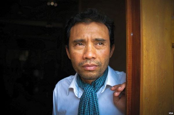 Man with short dark hair wearing a white shirt and blue checkered scarf, looking pensively out from a doorway.