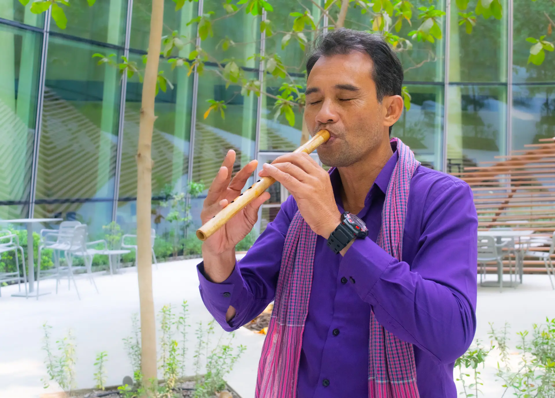 Man in a purple shirt playing a wooden flute outdoors near glass windows and greenery.