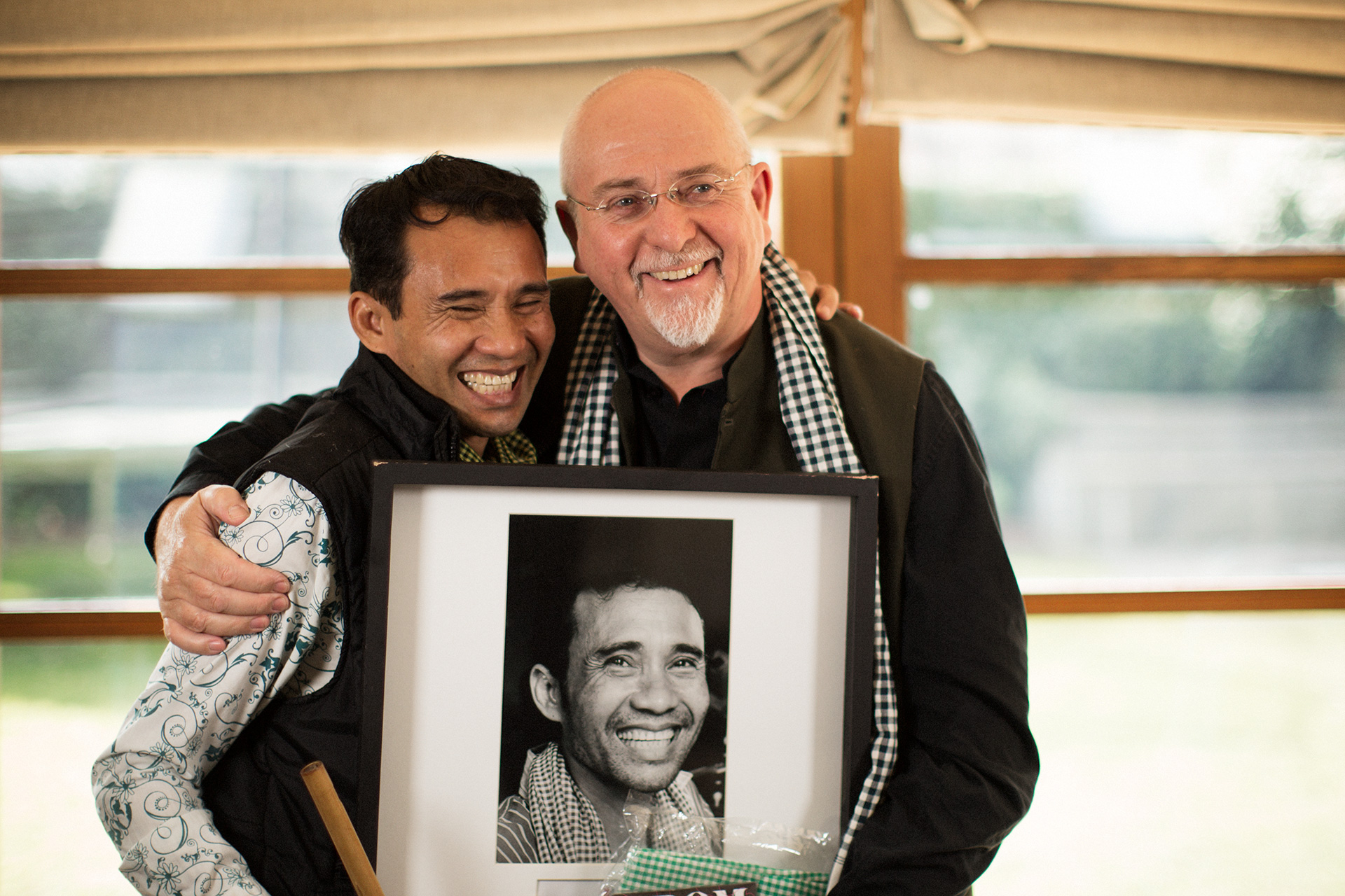 Two men smiling and hugging, one holding a framed black-and-white photo of the other smiling smiling.