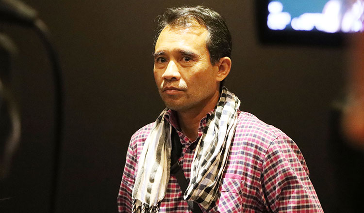 Man in a pink and black checkered shirt with a black and white scarf around his neck, standing against a dark background.