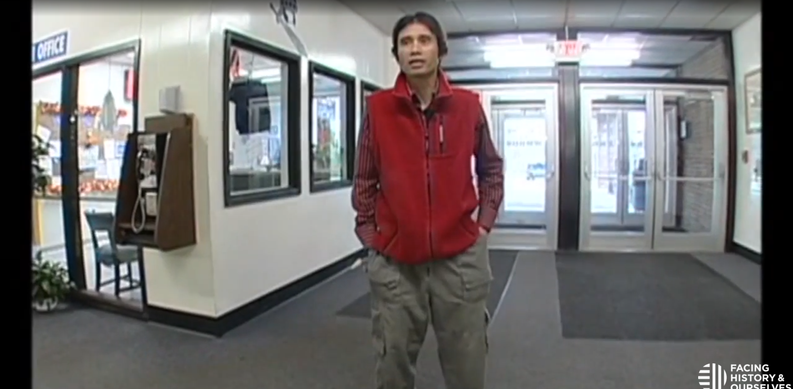 Man wearing a red vest and cargo pants standing in a hallway near an office entrance and glass doors.