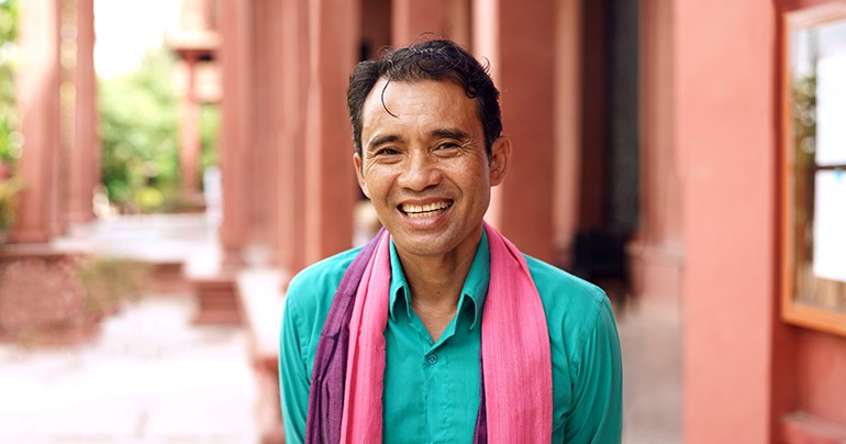 Smiling man wearing a teal shirt with pink and purple scarves standing outdoors near red buildings.