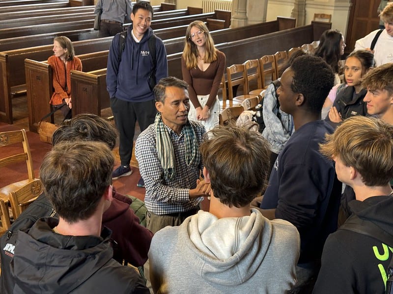 Man with a scarf speaking to a group of attentive young people inside a church or chapel with wooden pews.