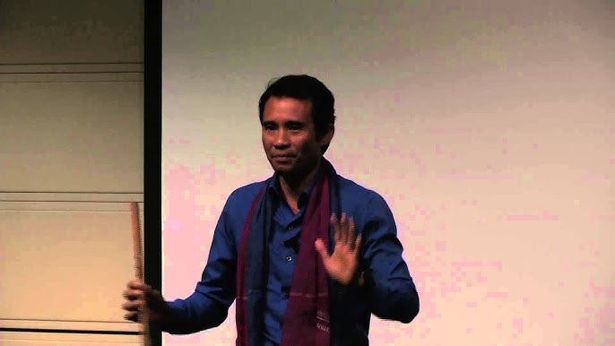 Man in blue shirt with maroon scarf holds a stick and gestures with his hand against a plain background.