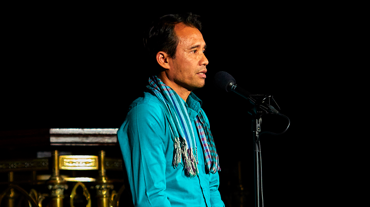 Man in teal shirt and striped scarf speaking into a microphone against a dark background.