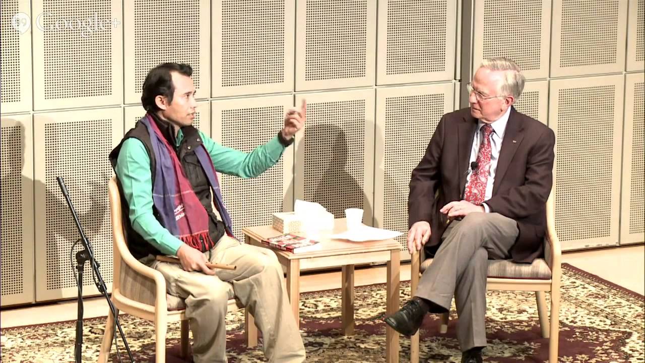Man in plaid shirt and glasses speaking at a podium with a microphone and a glass of water in front of him.