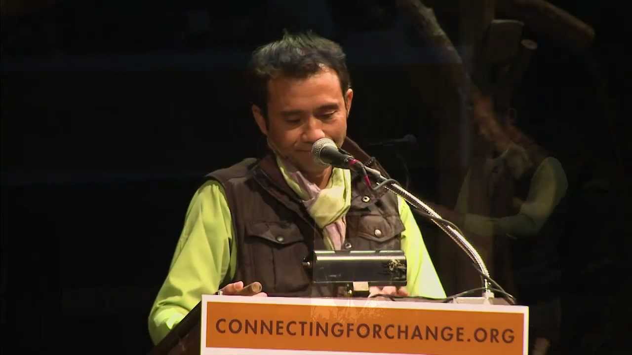 Man wearing glasses and a colorful checkered shirt speaking at a podium with a microphone and water bottle.