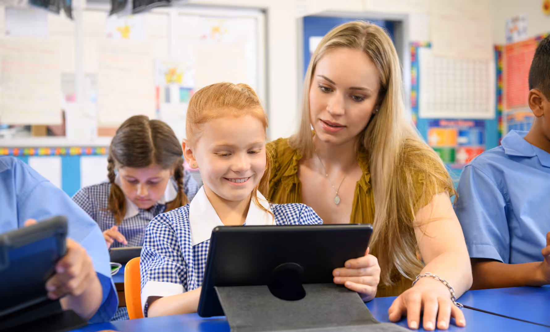 Primary school teacher helping students complete NAPLAN-style practice test on a tablet in the classroom.