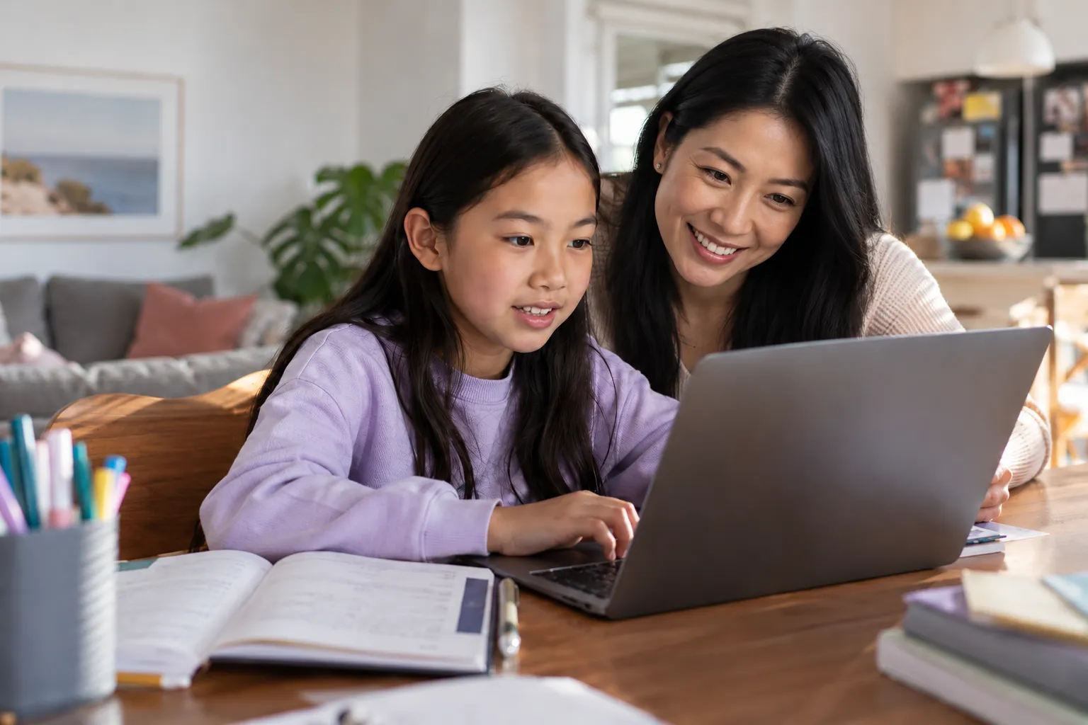 A mother and her daughter smiling as they use a laptop together at the kitchen table.