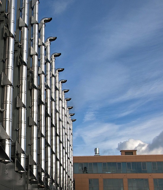 Modern building exterior with tall metallic pipes extending vertically against blue sky with clouds.