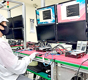 Technician in cleanroom suit testing multiple computer monitors with connected hardware on a lab bench.