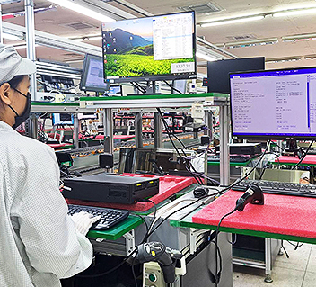 Worker in protective gear operating testing equipment with computer monitors in a factory setting.