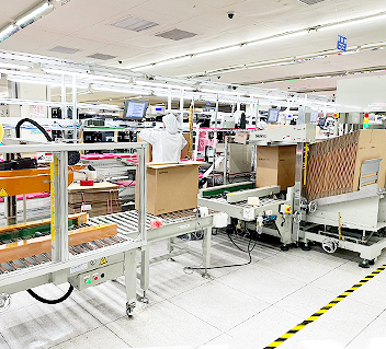 Production line worker handling cardboard boxes in a brightly lit factory with machinery and monitors.